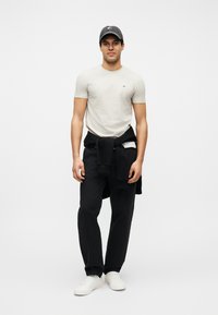 Νεαρός άνδρας που φοράει ανοιχτό γκρι μπλουζάκι, μαύρο παντελόνι, μαύρο φούτερ δεμένο γύρω από τη μέση, σκουρόχρωμο καπέλο και λευκά αθλητικά παπούτσια, στέκεται μπροστά σε λευκό φόντο.