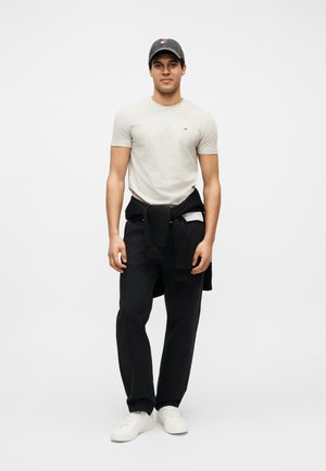 Mladi moški v svetlo sivi majici, črnih hlačah, črni jopa zavezani okoli pasu, temni kapi in belih supergah, stoji pred belim ozadjem.