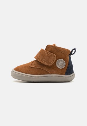 En alce marrone con cinturino in velcro, pannello posteriore blu navy, suola in gomma grigia e toppa rotonda con logo sul lato.