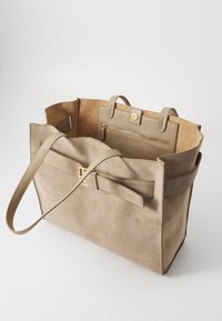 Sac cabas en daim beige avec deux poignées supérieures, poche intérieure zippée et détail nœud sur le devant. Texture lisse avec un design minimaliste.