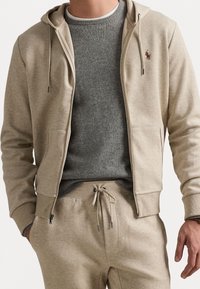 Beige huvtröja med dragkedja, dragsnören, ribbade muddar och framfickor över en grå crewneck-tröja, kombinerad med matchande mjukisbyxor.