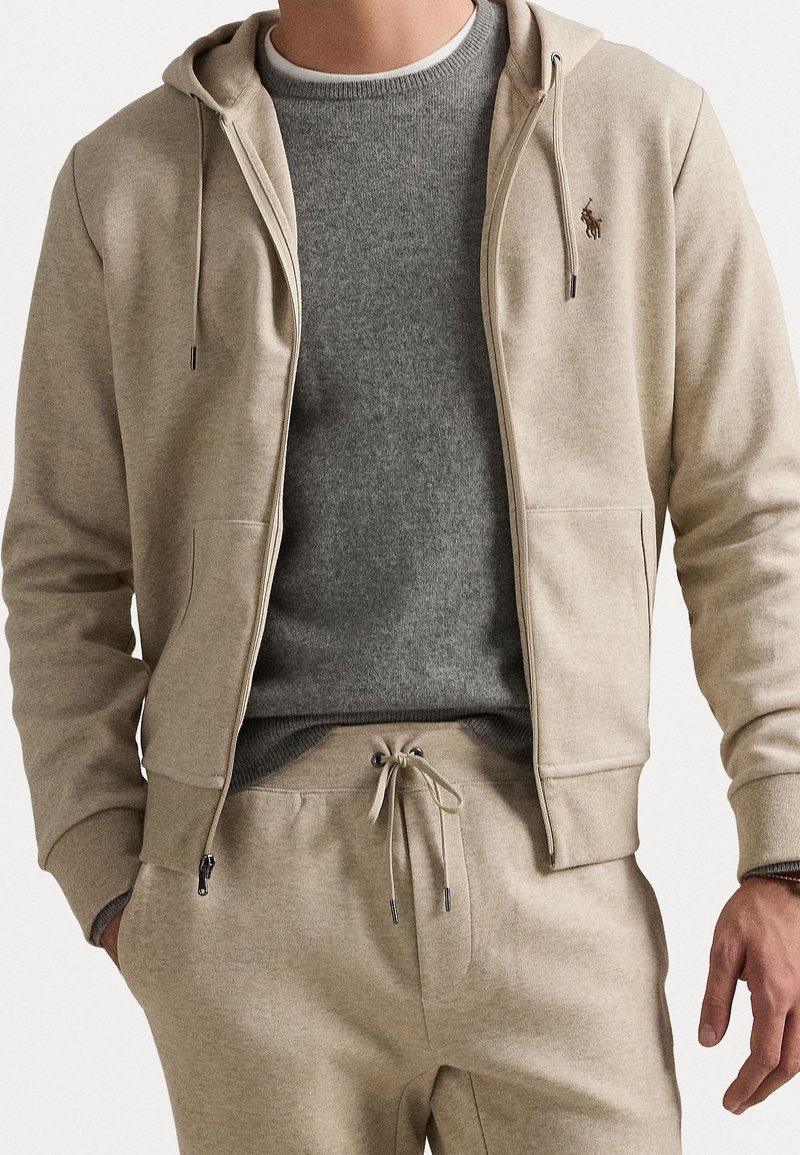 Beige huvtröja med dragkedja, dragsnören, ribbade muddar och framfickor över en grå crewneck-tröja, kombinerad med matchande mjukisbyxor.