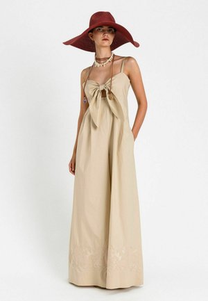 Donna che indossa una tuta beige a gamba larga con un grande fiocco frontale, cappello rosso a tesa larga e collana di conchiglie su uno sfondo bianco semplice.