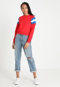 Sweatshirt rosso corto con strisce blu e bianche sulle maniche, abbinato a jeans blu chiaro strappati e sneakers bianche.