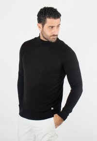 Pull noir à col montant à manches longues fabriqué dans un tissu lisse, doté d'un col et d'un ourlet côtelés, avec une petite étiquette logo en bas.