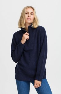 Námornícky pulóver na polovičný zips vyrobený z textúrovanej látky s uvoľneným strihom, s golierom na zips a bočnými rozparkom, kombinovaný s modrými džínami.