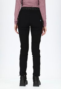 Black outdoor pants with a fitted design, tapered legs, zippered ankle cuffs, and a logo waistband, worn with sturdy black shoes.