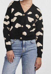 Cardigan noir avec de grands motifs en forme de cœur beige, col en V, et trois boutons marron. Porté par-dessus un jean bleu clair, avec une coupe décontractée.