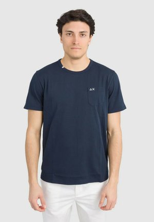 Giovane uomo con capelli scuri che indossa una t-shirt blu navy a maniche corte con un piccolo logo sulla tasca del petto e pantaloni bianchi.