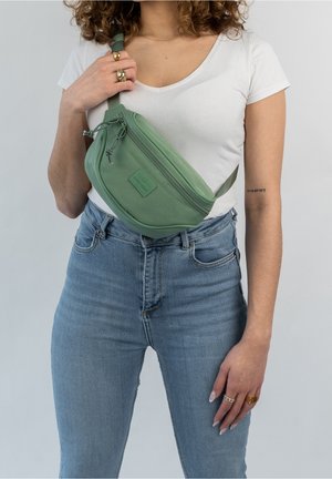 Femme portant un jean bleu clair et un t-shirt blanc avec un sac bandoulière vert et plusieurs bagues aux doigts sur un fond uni.