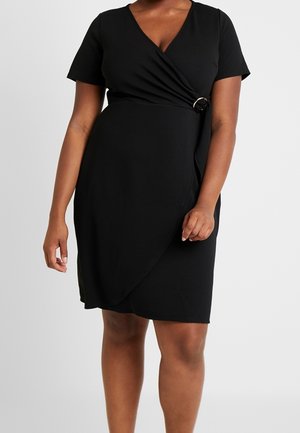 Vrouw draagt een zwarte wikkeljurk met korte mouwen en een zilveren ringdetail in de taille tegen een effen grijze achtergrond.