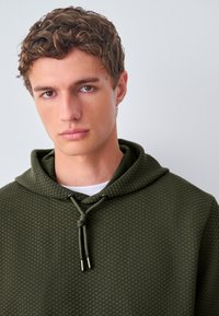 Sudadera con capucha verde caqui con un patrón texturizado de diamantes, capucha con cordón y puntas metálicas. Llevada sobre una camisa blanca.