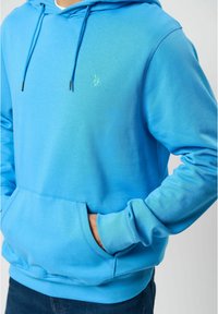 Světle modrá mikina s klokankou, kapucí se stahovací šňůrkou a jemně vyšívaným logem na hrudi. Měkká textura látky.