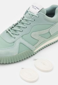 Zapatilla verde claro con parte superior de gamuza texturizada y cuero suave, con una suela gruesa y cordones blancos planos a un lado.