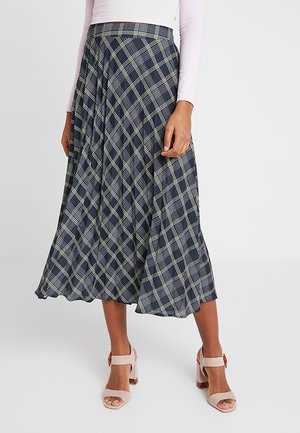 Marineblauer, plissierter Midirock mit weiß-grünem Karomuster, ausgestattet mit einem breiten Bund und fließendem Stoff. Kombiniert mit hellrosa Blockabsatzschuhen.