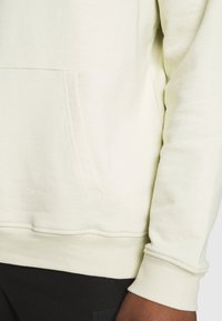 Felpa leggera color crema con tasca a marsupio, polsini e bordo a coste; realizzata in morbido materiale con dettagli di cucitura visibili.