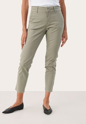 Pantalon slim-fit vert olive clair porté par une personne debout, associé à des chaussures plates noires pointues et un haut blanc.