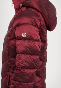 Burgundin värinen puhvitakki, jossa on kiiltävä pinta, sivuissa vetoketjulliset taskut ja huppu. Takissa on kultainen logovivahde hihassa.