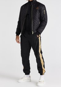 Svart vadderad bomberjacka med guldiga accenter, kombinerad med svarta träningsoveraller med guldiga sidostråkar. Bärs med vita sneakers.