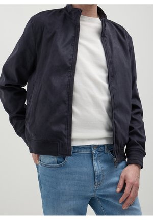 Homme portant une veste noire zippée, un pull blanc col rond et un jean bleu sur un fond clair uni.