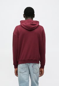 Kestenjasto smeđa majica s kapuljačom sa vezicama, rebrastim manšetama i rubom. Nosi se preko svijetloplavih traperica, prikazujući opušteni kroj i meku teksturu.