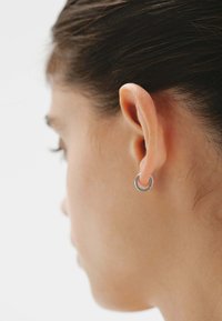 Pendiente de aro plateado mostrado en el lóbulo de la oreja, de forma circular con una superficie lisa y un brillo sutil, contrastando con un fondo neutro.