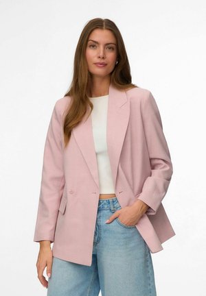Femme portant un blazer oversize rose clair, un haut court blanc et un jean bleu clair, debout avec une main dans la poche devant un fond blanc.