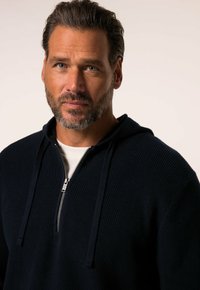Donkerblauwe ritssluiting hoodie met ribbelstructuur, capuchon met trekkoord en zilveren ritsdetails. Gezicht en bovenlichaam van het model zichtbaar.