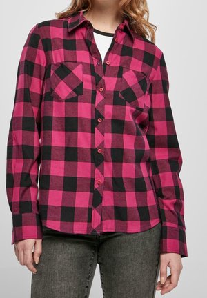 Kvinde iført en pink og sort ternet skjorte med knapper over en hvid top, kombineret med mørkegrå jeans, vist fra nakke til hofter.
