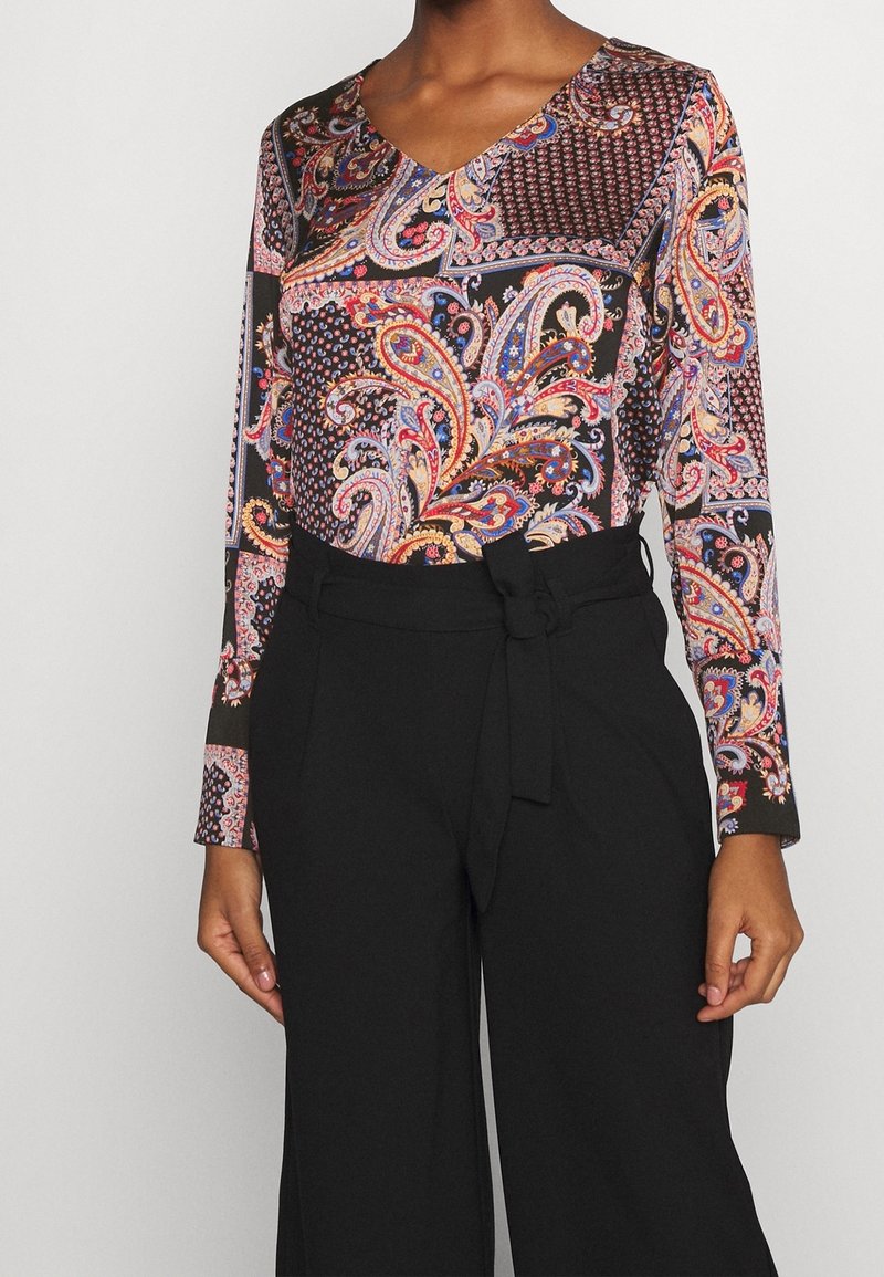Langärmliges Oberteil mit einem bunten Paisley-Muster auf schwarzem Hintergrund, kombiniert mit hochgeschnittenen schwarzen Hosen mit einem gebundenen Gürtel.