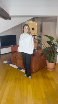 Vit sweatshirt med svart text, svarta byxor och silvriga skor. Brun manchestersoffa, grön växt, trägolv och TV i bakgrunden.