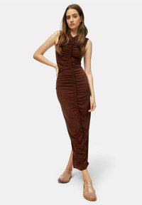Abito maxi senza maniche marrone con dettagli arricciati lungo la parte anteriore e i lati, realizzato in tessuto elasticizzato. La modella indossa sandali trasparenti.
