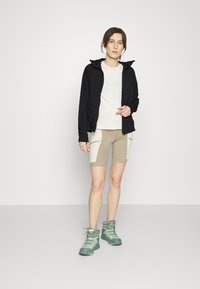 Sort letvægtsjakke, creme T-shirt og beige cykshorts med hvide paneldetaljer, parret med teal vandrestøvler med gule snørebånd.