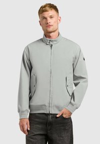 Jeune homme portant une veste zippée gris clair à col montant et poches à rabat, associée à un jean foncé, debout devant un fond uni.