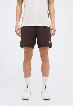 Mężczyzna ubrany w brązowe szorty sportowe, białe skarpetki, białe trampki i biały sportowy t-shirt z kolorowymi paskami po bokach, stojący na jednolitym tle.