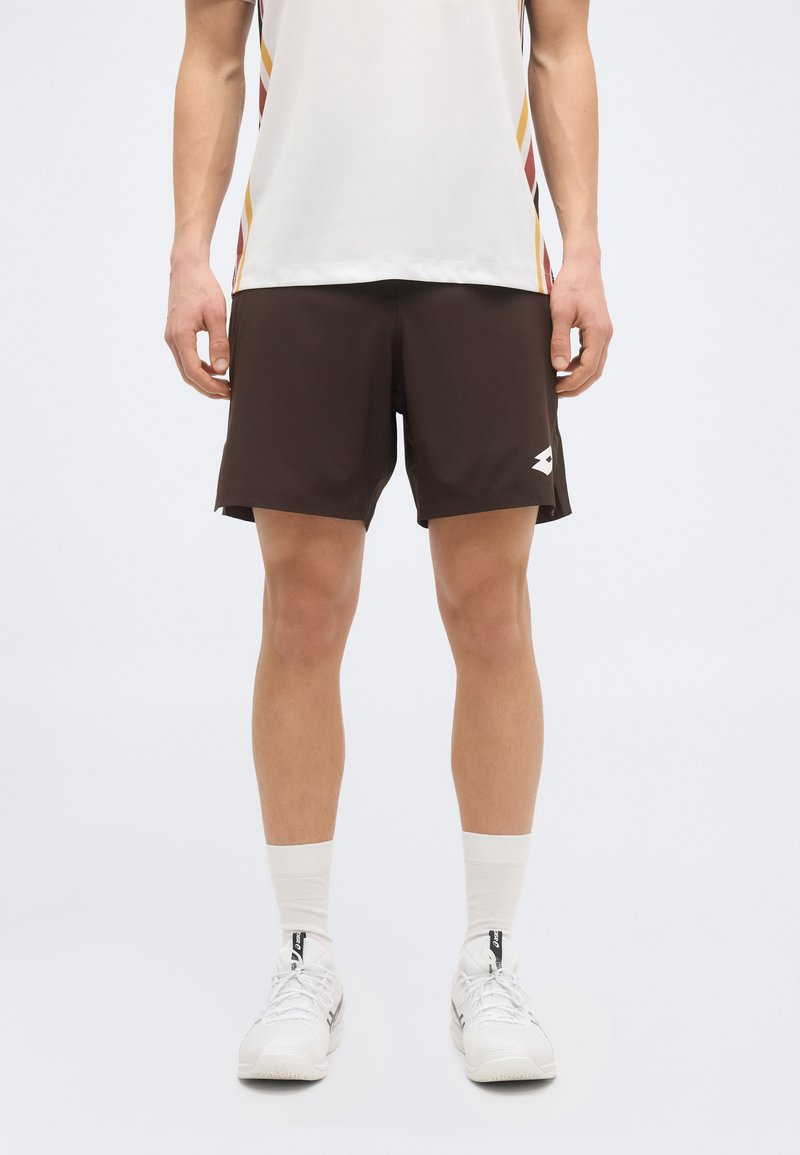 Άνδρας που φορά καφέ αθλητικό σορτς, λευκές κάλτσες, λευκά αθλητικά παπούτσια και λευκό αθλητικό μπλουζάκι με πολύχρωμες ρίγες στο πλάι, στέκεται μπροστά σε ένα απλό φόντο.