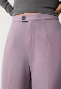 Pantalons lavande en tissu lisse, avec fermeture à un bouton et plis sur le devant. La taille est structurée avec un design épuré.