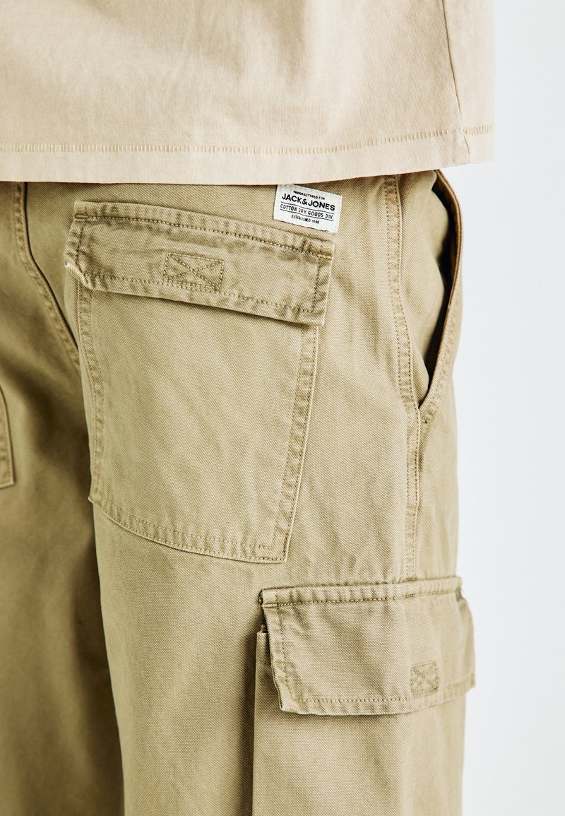 Pantalones cargo color beige con múltiples bolsillos tipo parche, detalles de costura y una etiqueta de tela con el nombre de la marca en la cintura.