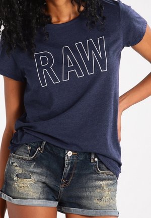 Kvinde iført navyblå t-shirt med teksten "RAW" og slidte denimshorts, hånd hvilende på hoften, mod en ensfarvet baggrund.