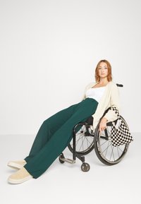 Junge Frau sitzt im Rollstuhl, trägt grüne Hose, weißes Top, cremefarbenen Cardigan und hält eine schwarz-weiß karierte Tasche.