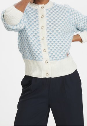 Kobieta ubrana w fakturowany, jasnoniebiesko-biały rozpinany kardigan z guzikami przypominającymi perły i luźne, ciemnogranatowe spodnie.
