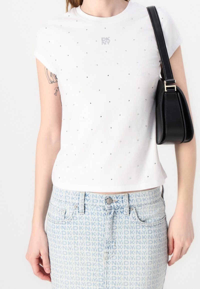 Personne portant une chemise blanche à manches courtes avec de petits strass et une jupe bleu clair ornée du logo MDKNY répété, tenant un sac à épaule noir.