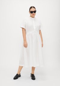 Weißes Hemdkleid mit kurzen Ärmeln, Knopfleiste, elastischem Bund und Seitentaschen; getragen mit schwarzen Loafers und übergroßen schwarzen Sonnenbrillen.