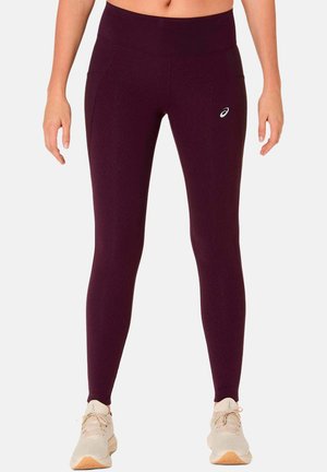 Donna che indossa leggings a vita alta di colore viola scuro con tasche laterali e scarpe sportive chiare su sfondo bianco.