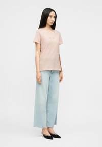 Μια γυναίκα στέκεται φορώντας ένα ανοιχτό ροζ T-shirt A|X, ανοιχτό μπλε παντελόνι καμπάνα κομμένο πάνω από τον αστράγαλο με ξεφτισμένα τελειώματα και μαύρα μυτερά παπούτσια slingback.