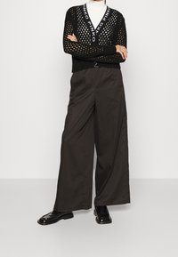 Svart stickad tröja med stort öppet nätmönster, V-ringning och logotypdetaljer. Kombinerad med vida, mörkbruna byxor och polerade skor.