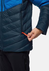 Giacca puffa in blu navy scuro e blu con trapuntatura a chevron, dotata di una tasca laterale con zip e un cordino arancione con l'etichetta "PACK AWAY".