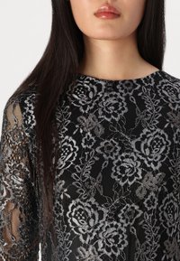Haut en dentelle noire avec une broderie florale argentée complexe et un col rond, des manches longues transparentes et un corps texturé.
