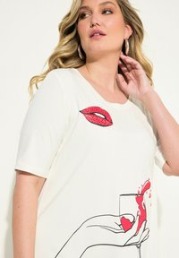 Vit t-shirt med kort ärm som har en grafisk design med röda läppar och en hand som håller ett glas med röda vätskesplashes. Mjuk tyg, avslappnad passform.