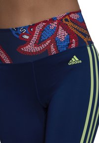 Zbliżenie osoby w granatowych legginsach treningowych Adidas z neonowymi zielonymi paskami i kolorowym wzorzastym pasem.